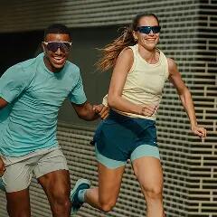 Ein Mann und eine Frau joggen mit freudigem Lächeln nebeneinander her. Beide tragen sportliche Sonnenbrillen und funktionale Laufbekleidung – er ein hellblaues T-Shirt und graue Shorts, sie ein cremefarbenes Tanktop und dunkelblaue Doppel-Shorts. Der Hintergrund zeigt eine moderne Fassade mit dunklen Fliesen, was die Dynamik der Bewegung unterstreicht. Die Szene strahlt Energie, Spaß am Sport und Gemeinschaft aus.