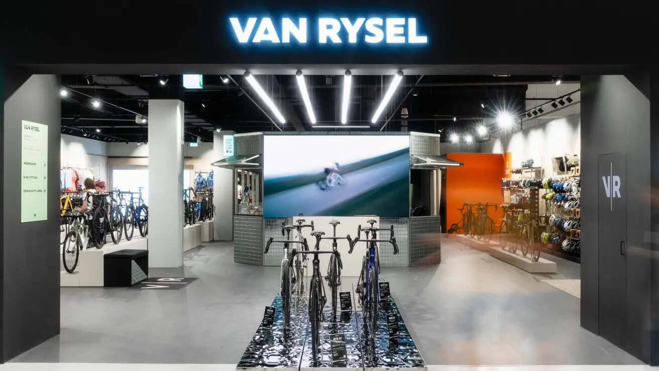 Die Außenansicht eines modernen VAN RYSEL Fahrradgeschäfts, betrachtet von der Eingangstür aus. Über dem Eingang leuchtet ein auffälliger, neonblauer „VAN RYSEL“-Schriftzug. Das Innere des Geschäfts ist hell beleuchtet und modern gestaltet. Im Mittelpunkt stehen drei perfekt präsentierte Rennräder auf einem Podest. Dahinter befindet sich ein Tresen mit einem großen Bildschirm, der ein Video von einem Radfahrer in Action zeigt. Zur Linken sind Kleiderständer mit Radsportbekleidung und weitere ausgestellte Fahrräder an einer Wand mit dem Text „ON ROAD“ zu sehen. Auf der rechten Seite gibt es ein Regal voller bunter Helme und eine dunkle Tür mit dem diskreten „VR“-Logo. Der Laden wirkt premium und gut sortiert, mit Sichtbetonboden und moderner Deckenbeleuchtung.
