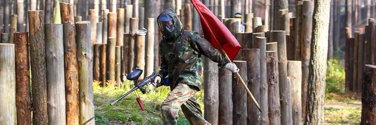 Mężczyzna biegajacy z pistoletem od paintballa i flagą w ręku grający w grę zdobywanie flagi