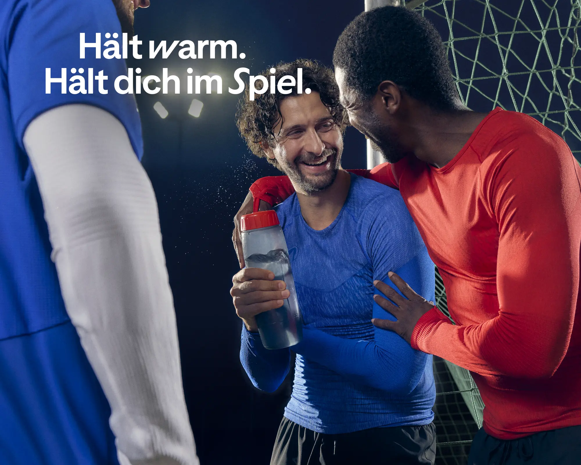 Drei lachende Sportler stehen in Funktionskleidung zusammen am Spielfeldrand, während oben links der Text „Hält warm. Hält dich im Spiel.“ eingeblendet ist.