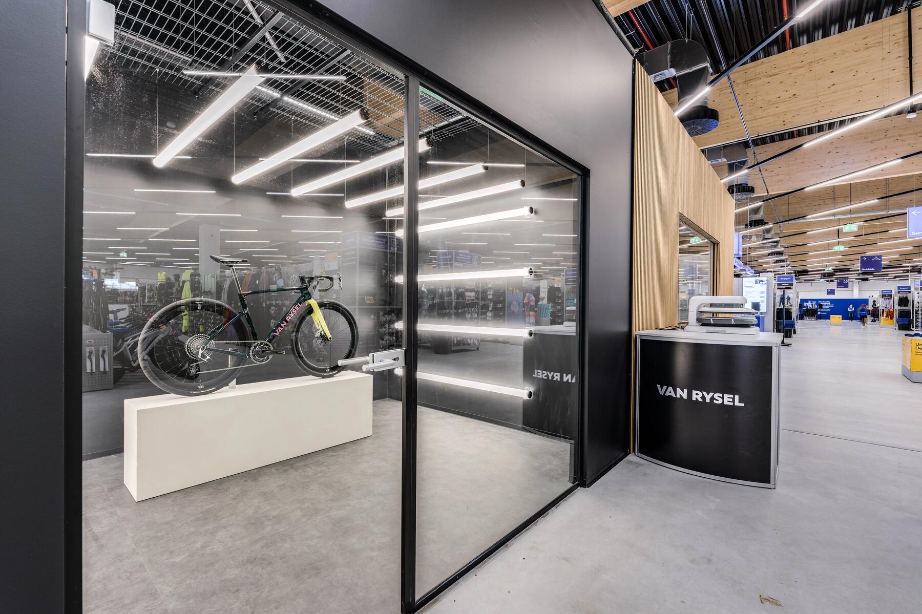 Dunkler Raum mit Fahrradtrainer, graue Wände, reflektierende Metallstäbe, Flachbildschirm, moderne Beleuchtung.