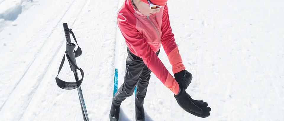 osoba w odzieży narciarskiej zakładająca rękawiczki narciarskie stojąc na nartach biegowych