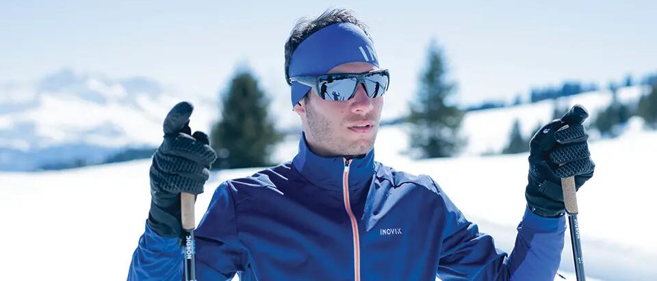 mężczyzna stojący w odzieży i okularach narciarskich trzymając kije narciarskie