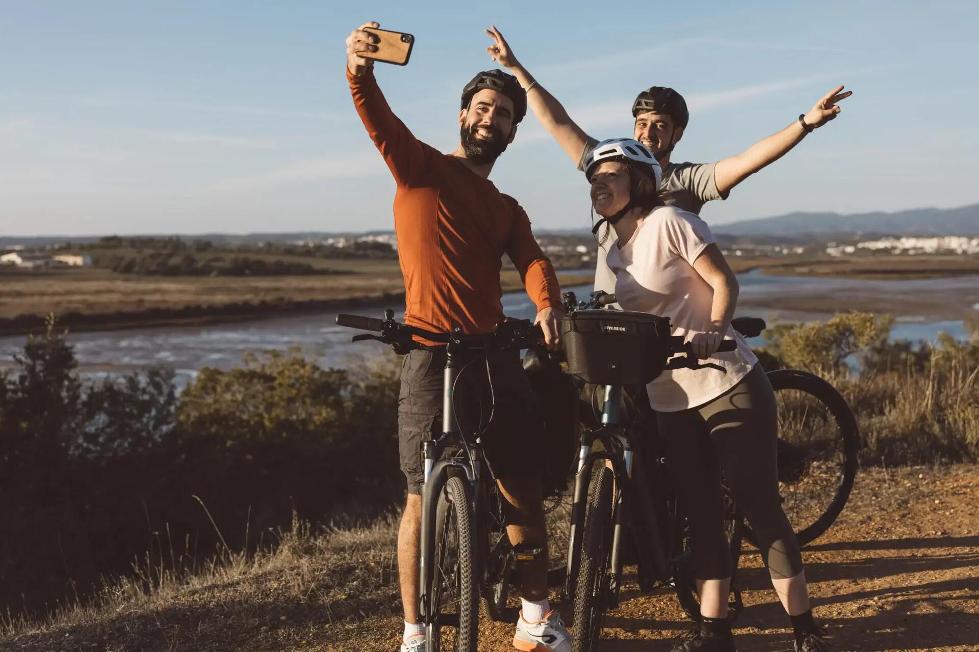 Ludzie w strojach sportowych i kaskach rowerowych  robiący sobie selfie w trakcie wyprawy rowerowej na rowerach elektrycznych  na tle rzeki