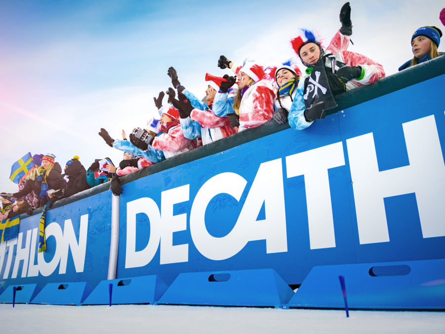 Franska supporters står vid en decathlon banner. Bannern avgränsar åskådarna från skidspåret.