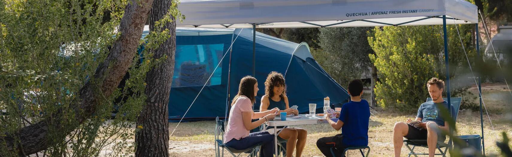 a family sat in a campsite,  the woman and two children are playing a game of cards and the man is sat reading 