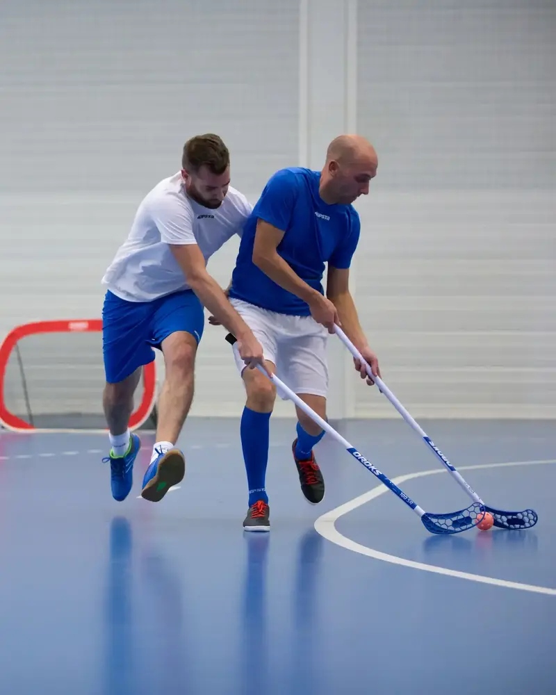 Drei Männer spielen intensiv Unihockey in einer Sporthalle, wobei ein Spieler in Weiß versucht, einem Gegner in Blau den Ball abzunehmen.