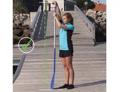 A girl on the pier with her paddle next to her reaching up to the top of the paddle to the correct hight.