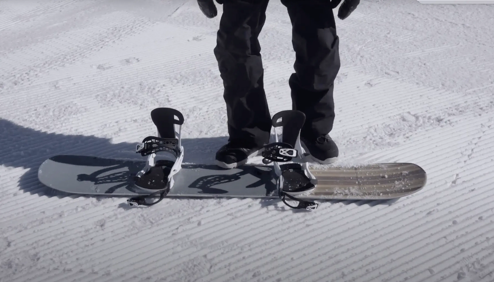Osoba stojąca obok leżącej na śniegu deski snowboardowej z przypiętymi wiązananiami snowboardowymi