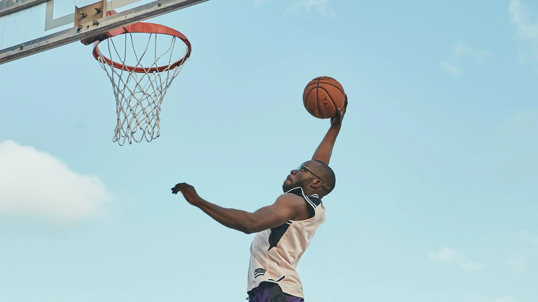 Basketballspieler beim Dunk, blauer Himmel, dynamische Bewegung, orangefarbener Ball, weißes Trikot, metallener Basketballkorb.