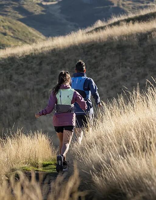 Zwei Personen joggen auf schmalem Weg durch goldgelbes, hohes Gras in hügeliger Landschaft bei Sonnenlicht.